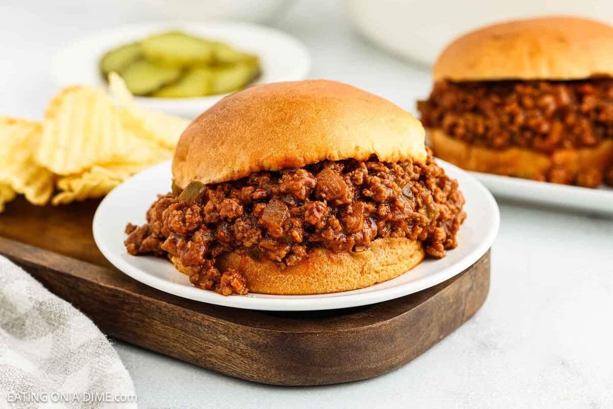 A classic Sloppy Joes sandwich with seasoned ground beef filling on a bun, served on a white plate with potato chips and pickles in the background—perfect for any Manwich craving.