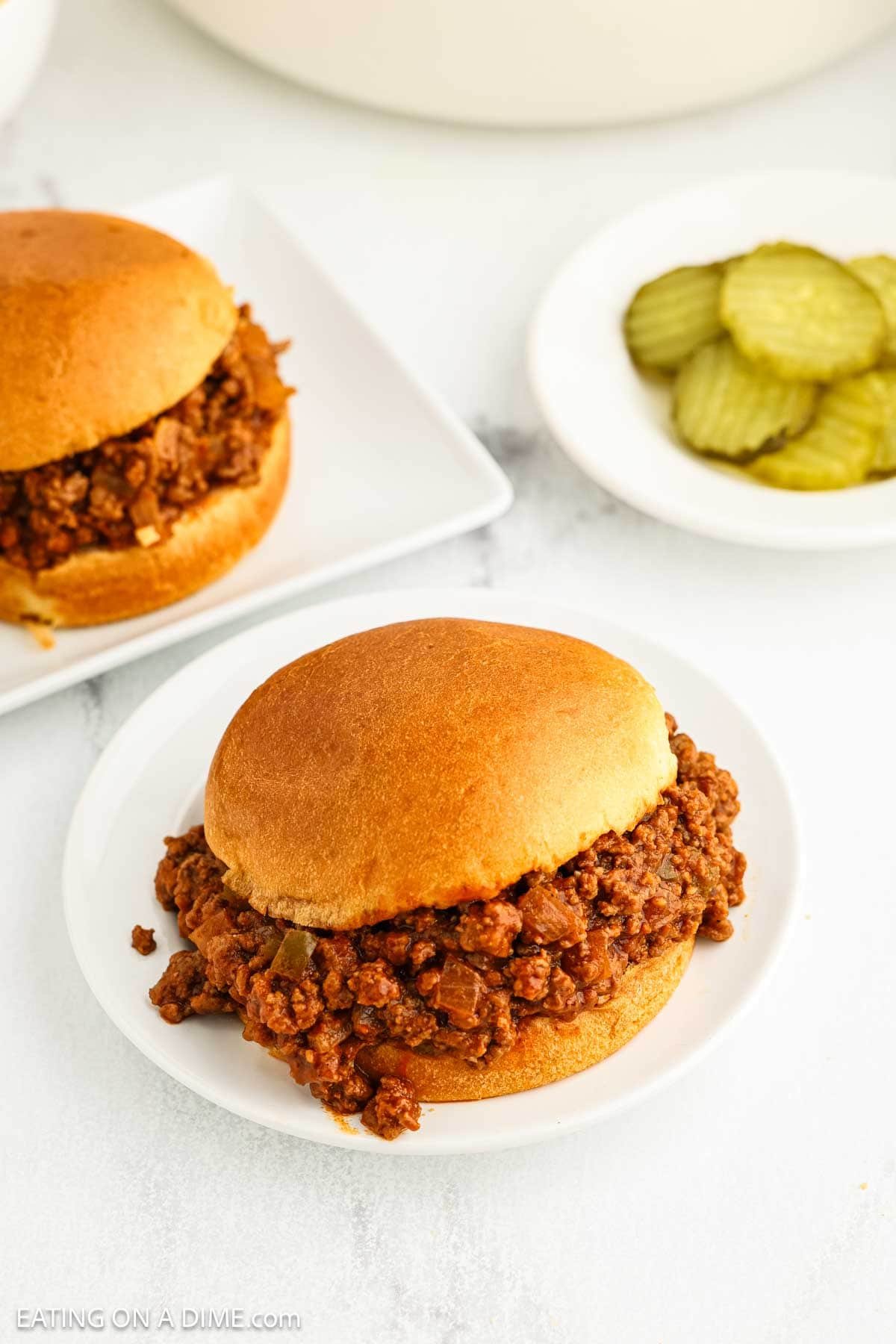 A classic Sloppy Joes sandwich with seasoned ground beef filling on a white plate, next to crisp pickle slices and another Manwich sloppy joe sandwich in the background.