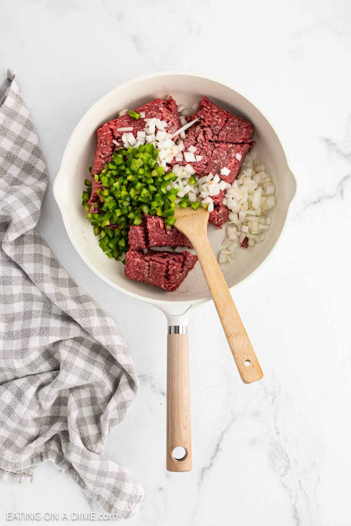 A white skillet contains raw ground beef, chopped green bell pepper, and diced onion—perfect beginnings for homemade Sloppy Joes or a tasty Manwhich. A wooden spoon rests in the skillet beside a gray and white checkered towel on the marble countertop.