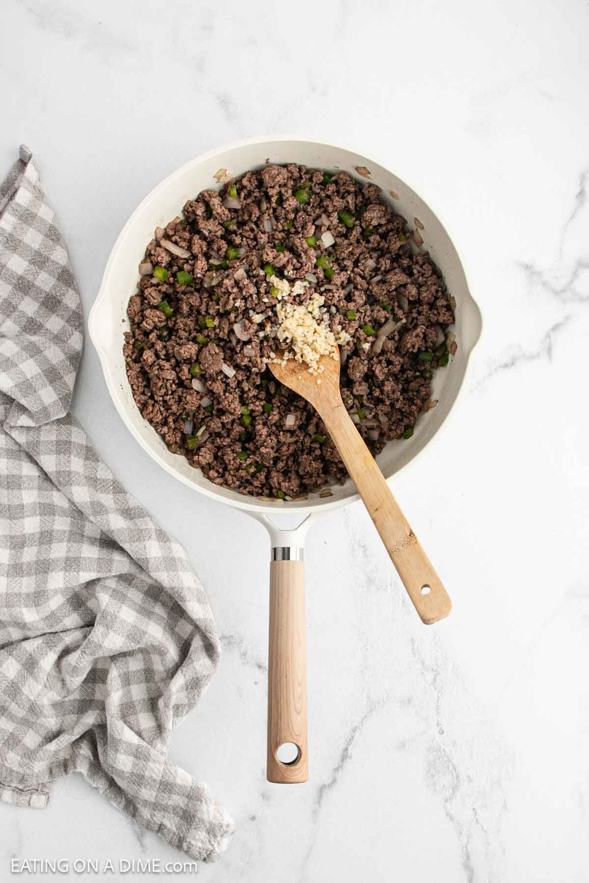A skillet filled with cooked ground beef, chopped green peppers, and onions—perfect for making Sloppy Joes. A wooden spoon holds minced garlic, while a gray and white checkered cloth rests beside the pan on a white marble surface.