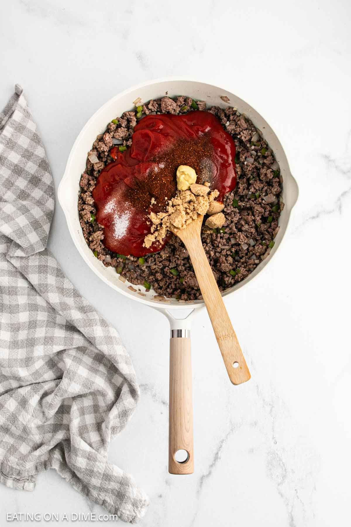 A skillet with cooked ground beef and vegetables, topped with ketchup, mustard, brown sugar, and spices—this Sloppy Joe recipe is stirred with a wooden spoon. A gray and white checkered towel sits beside the pan on a white countertop.