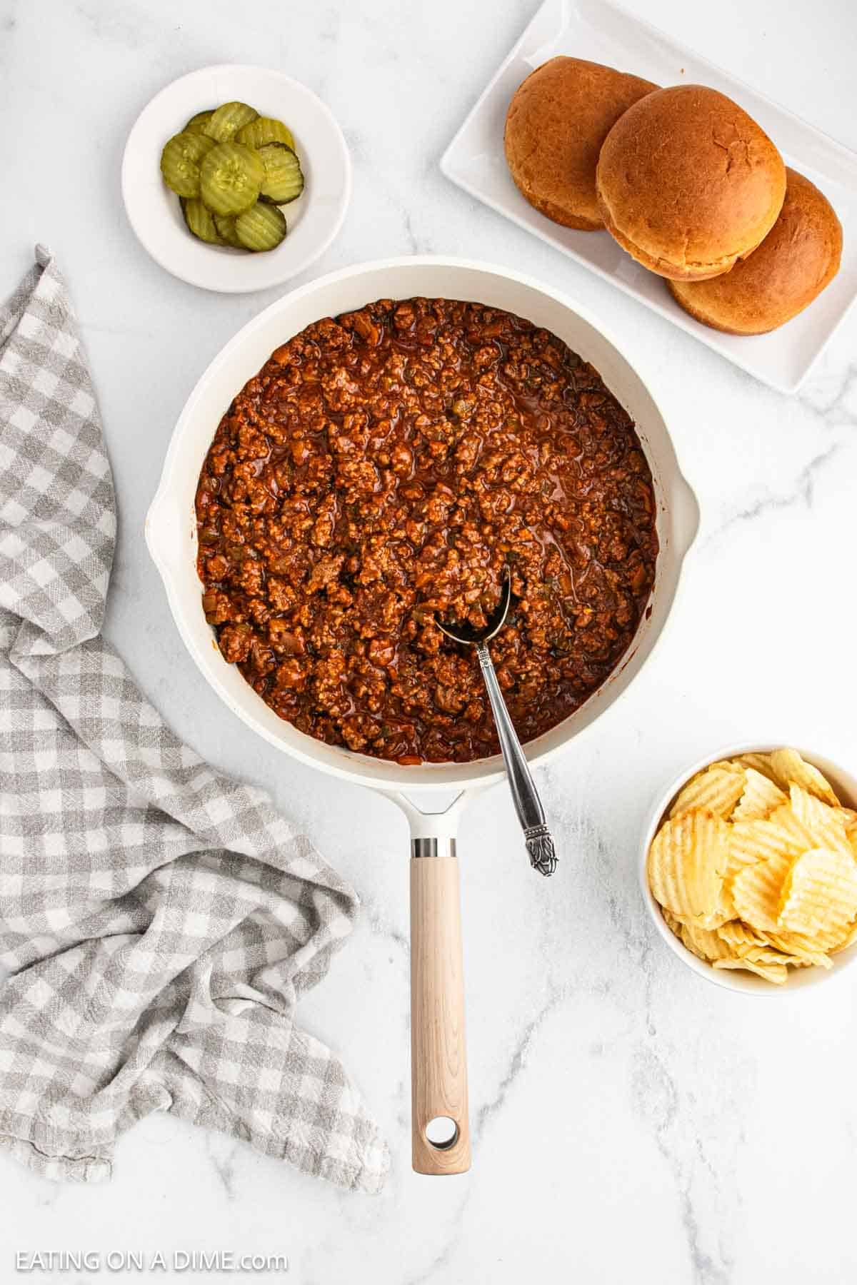 A skillet filled with savory Sloppy Joes meat and a spoon sits on a white countertop. Nearby are a plate with hamburger buns, a bowl of pickles, potato chips, and a gray checked napkin, ready for the perfect Manwhich meal.