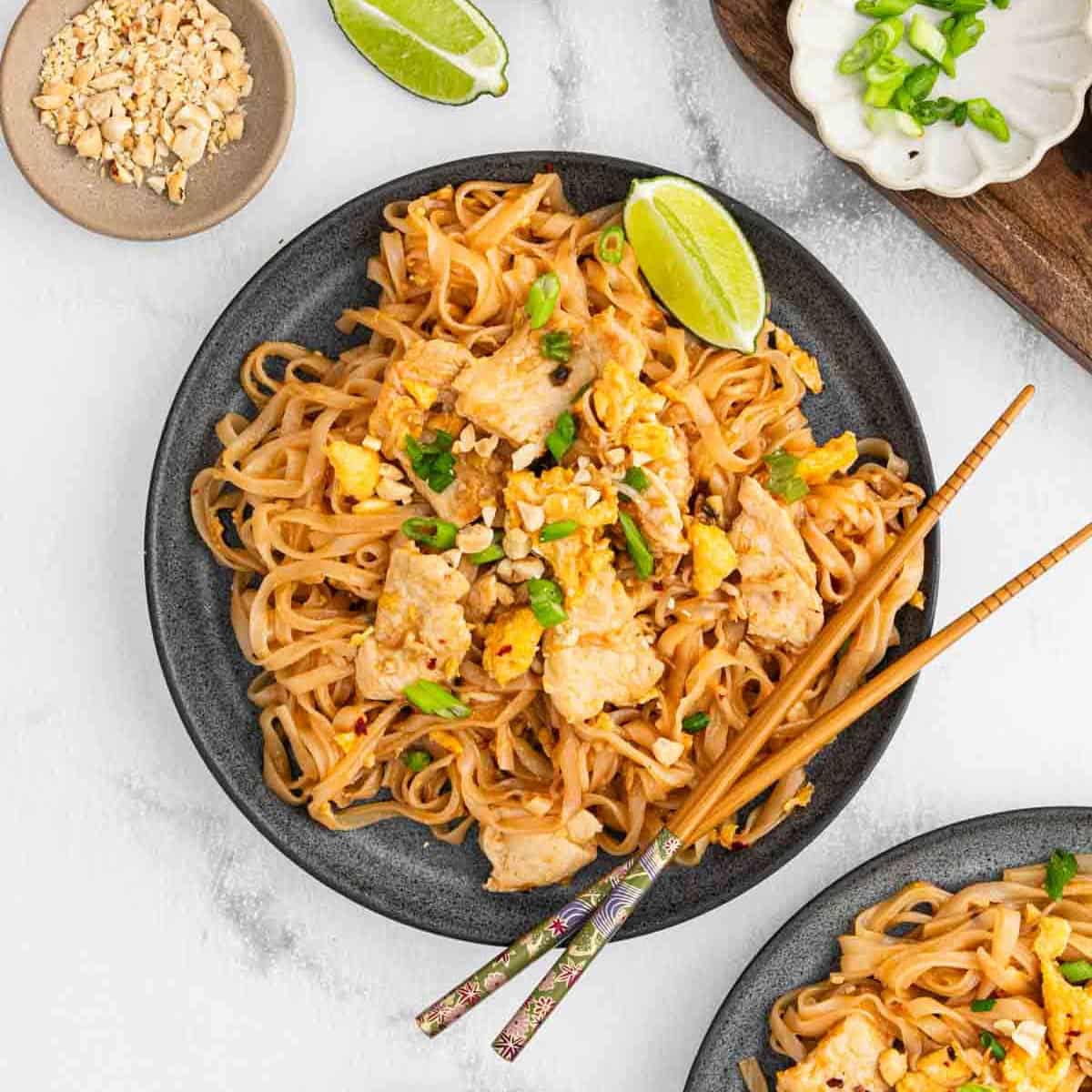 A plate of Pad Thai with tofu, garnished with chopped peanuts, green onions, and lime wedges. Chopsticks rest on the side of the plate, and extra peanuts and green onions are in small dishes nearby.