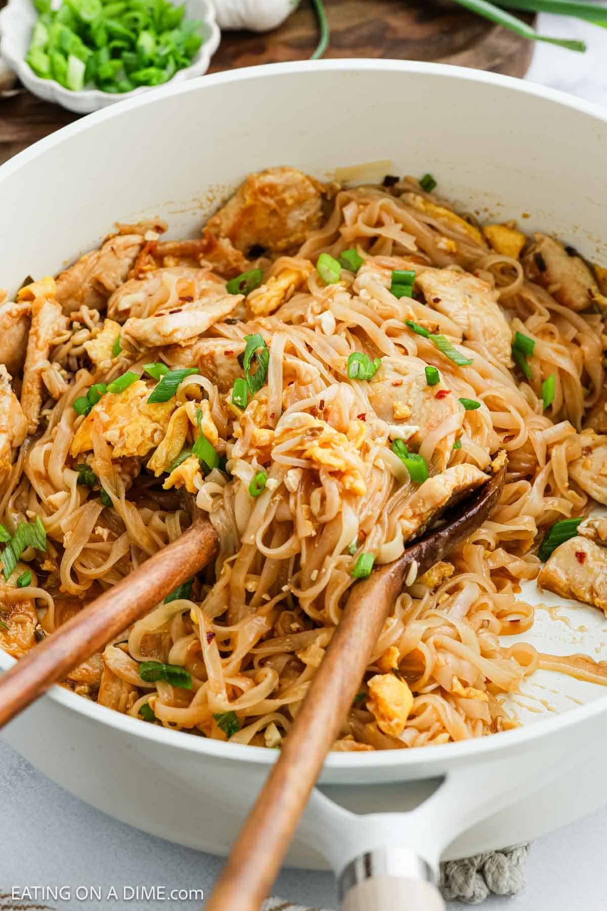 A skillet filled with stir-fried noodles, chicken pieces, scrambled eggs, and chopped green onions. Two wooden utensils are mixing the dish, and a bowl of sliced green onions is in the background.