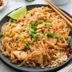 A plate of Pad Thai topped with chopped green onions, crushed peanuts, and chili flakes, garnished with a lime wedge and served with chopsticks. The flavorful Pad Thai features noodles, chicken, and scrambled egg.