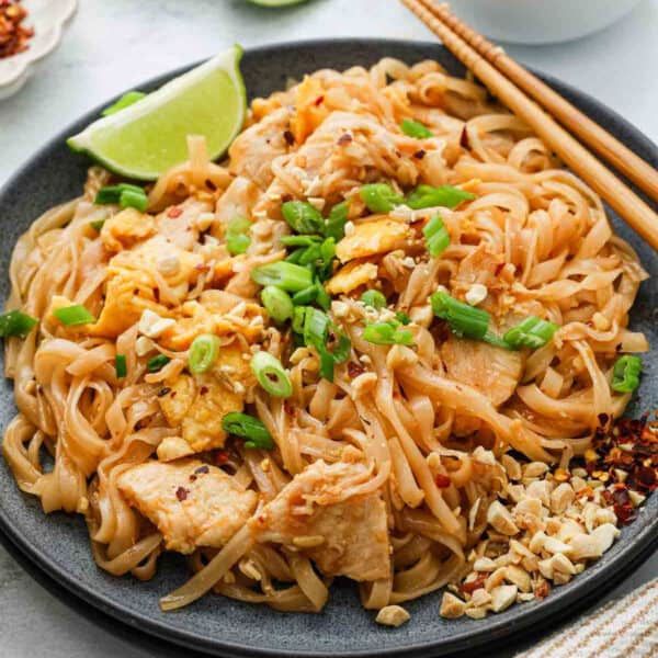 A plate of Pad Thai topped with chopped green onions, crushed peanuts, and chili flakes, garnished with a lime wedge and served with chopsticks. The flavorful Pad Thai features noodles, chicken, and scrambled egg.