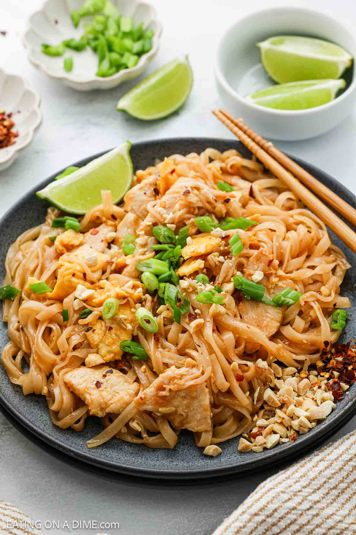 A plate of pad thai with chicken, garnished with chopped green onions, crushed peanuts, and lime wedges. Chopsticks rest on the side, and small bowls with more toppings are in the background.