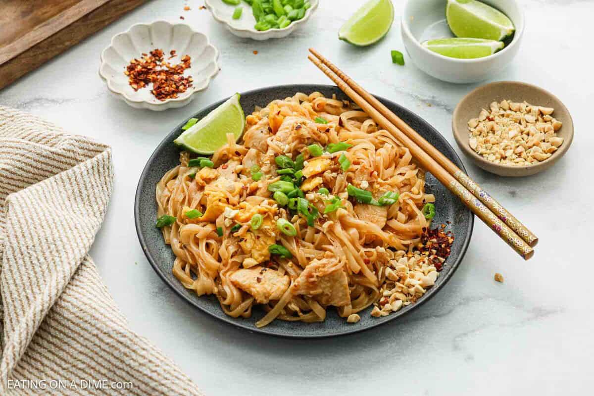 A plate of chicken pad Thai topped with chopped peanuts, green onions, and a lime wedge, with chopsticks resting on the plate. Nearby are bowls of crushed peanuts, chili flakes, and sliced green onions.