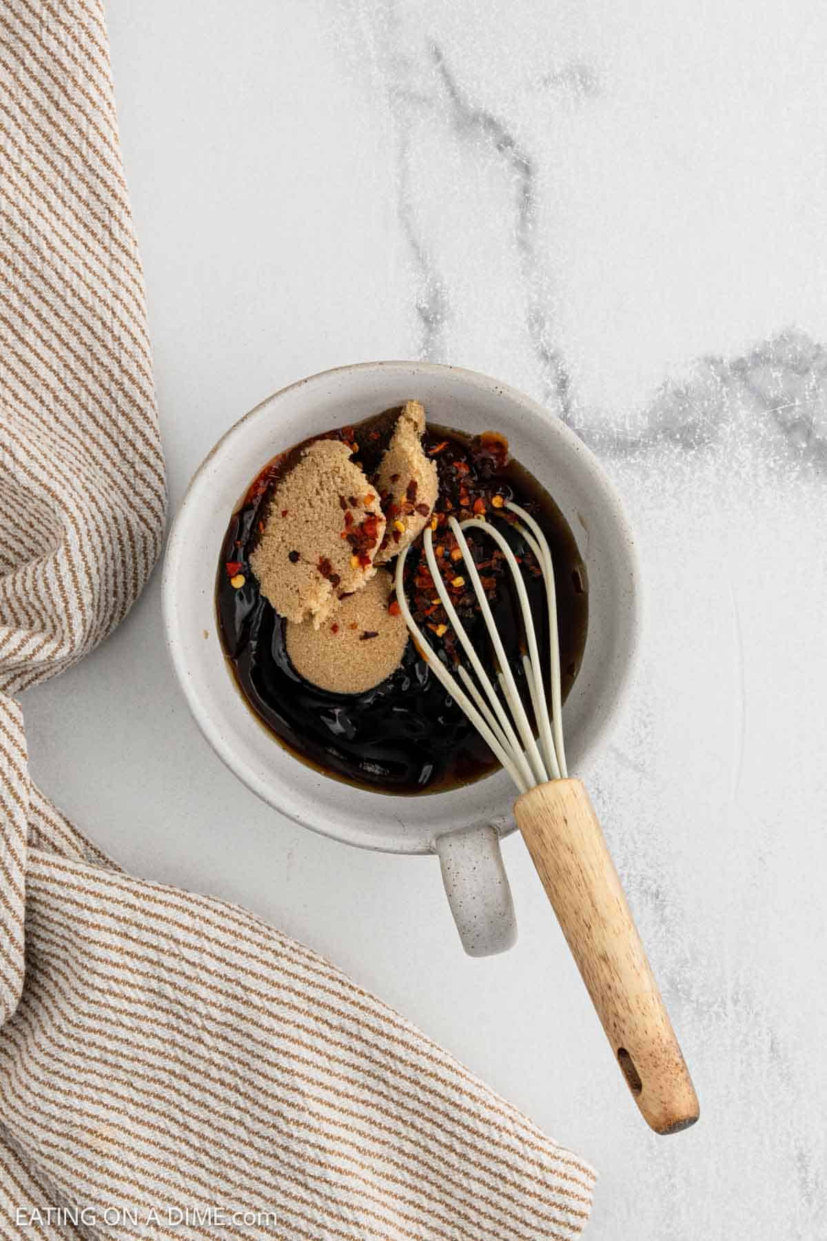 A bowl containing soy sauce, brown sugar, and red pepper flakes is being whisked with a wooden-handled whisk. A beige striped towel is placed next to the bowl on a white marble surface.