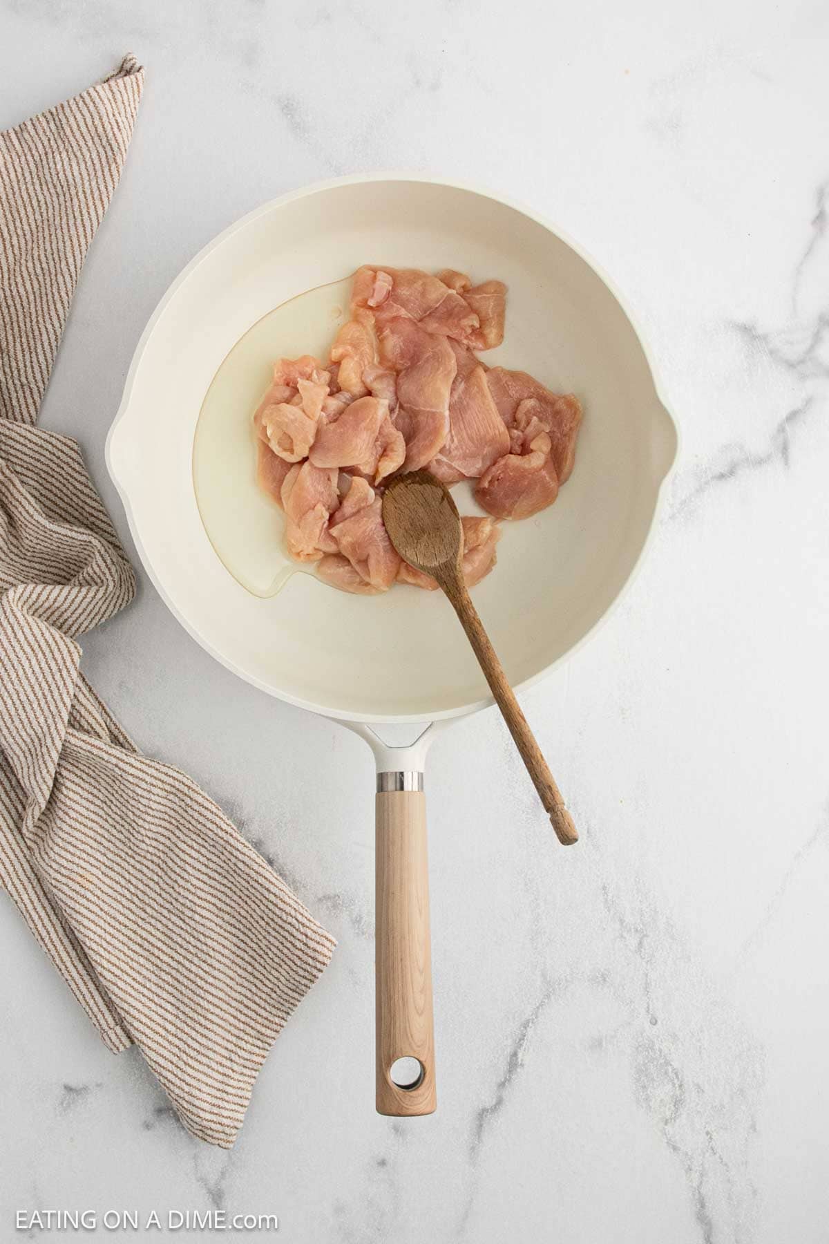 Raw pieces of chicken placed in a white skillet with a wooden spoon on top, next to a striped kitchen towel on a light marble surface.