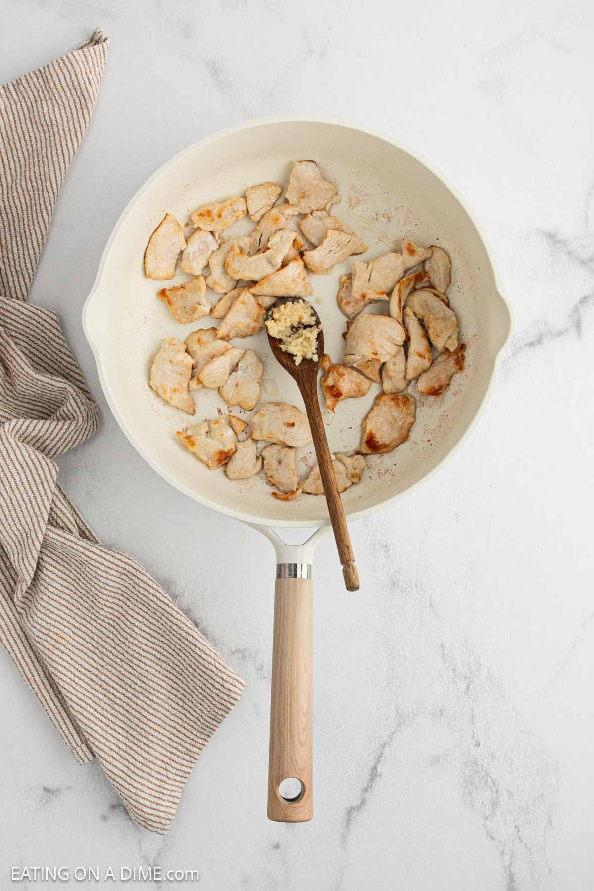 A white skillet with cooked sliced chicken and a wooden spoon holding minced garlic, on a marble surface next to a folded beige striped kitchen towel.