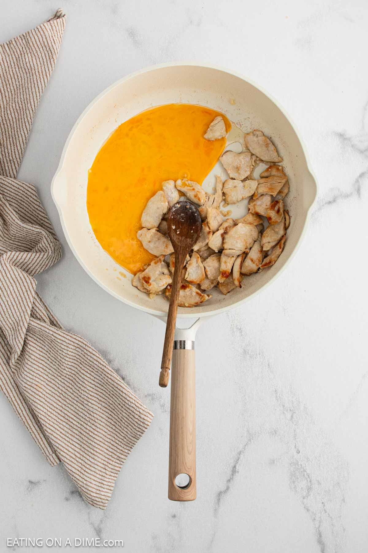 A white skillet contains cooked chicken pieces and beaten eggs, partially mixed together. A wooden spoon rests in the pan, and a beige-striped towel is nearby on a white marble surface.