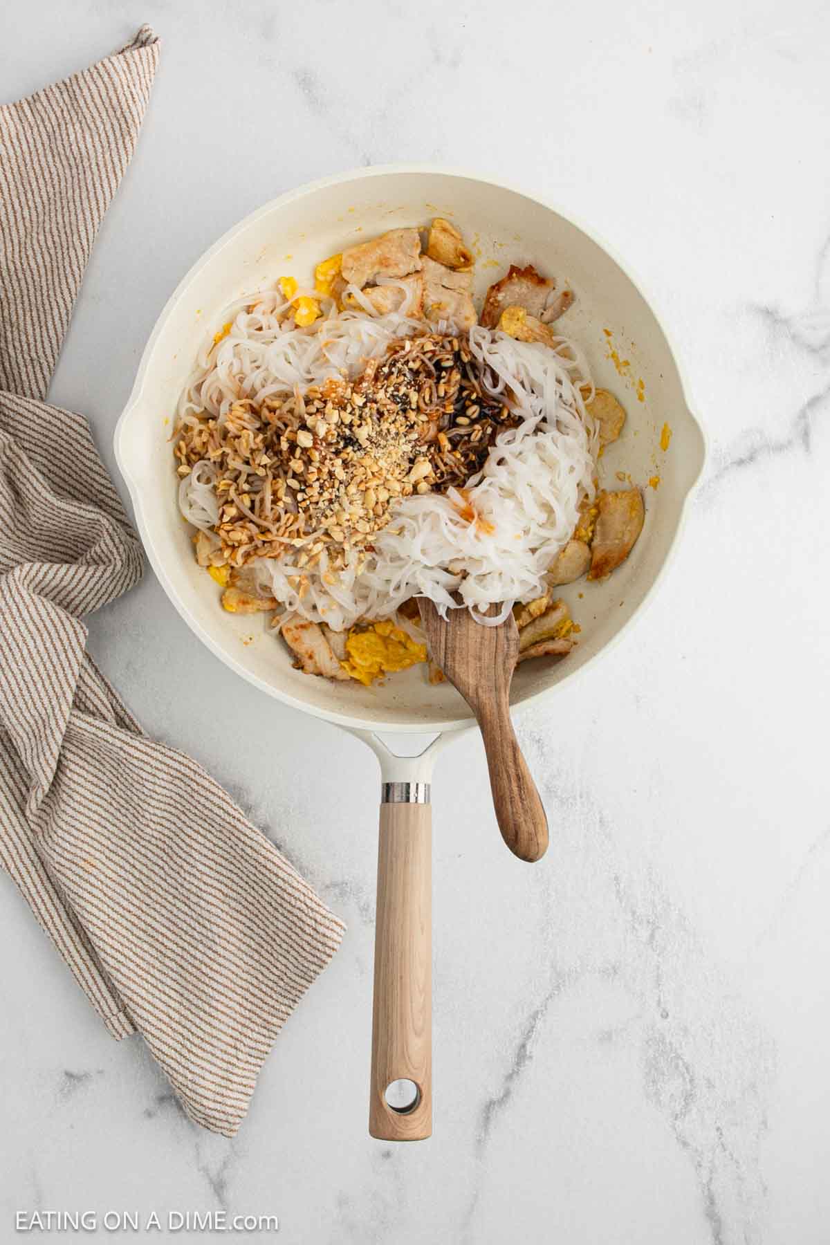 A skillet on a marble counter contains rice noodles, cooked chicken pieces, crushed peanuts, and sauce. A wooden spoon rests in the pan, and a striped kitchen towel is nearby.