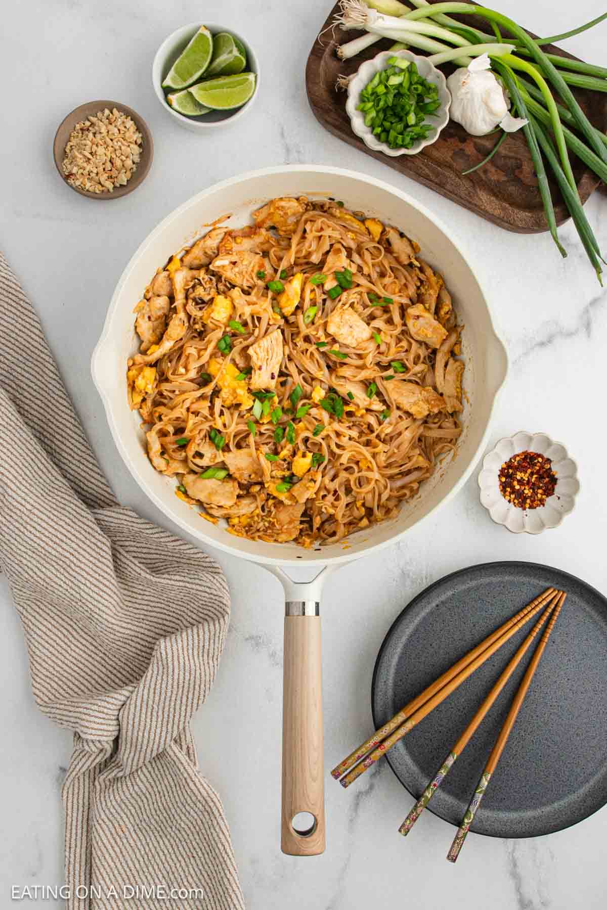 A skillet of chicken pad Thai with noodles, egg, and green onions sits on a white surface, surrounded by plates, chopsticks, lime wedges, chopped peanuts, green onions, and a striped napkin.