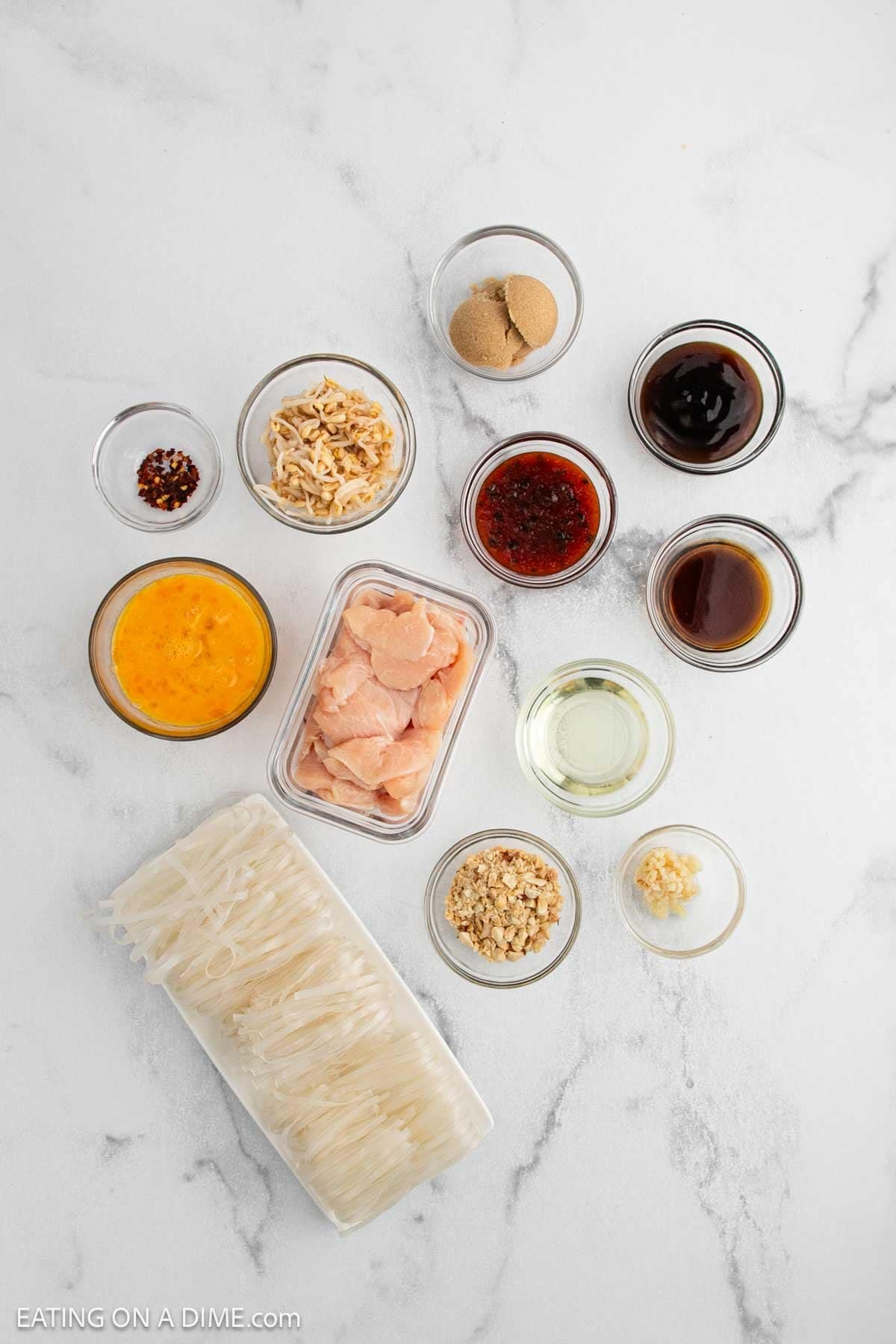 Various ingredients for a noodle dish are arranged on a white marble surface, including raw chicken pieces, uncooked rice noodles, beaten eggs, sauces, spices, sugar, chopped peanuts, and minced garlic in small bowls.