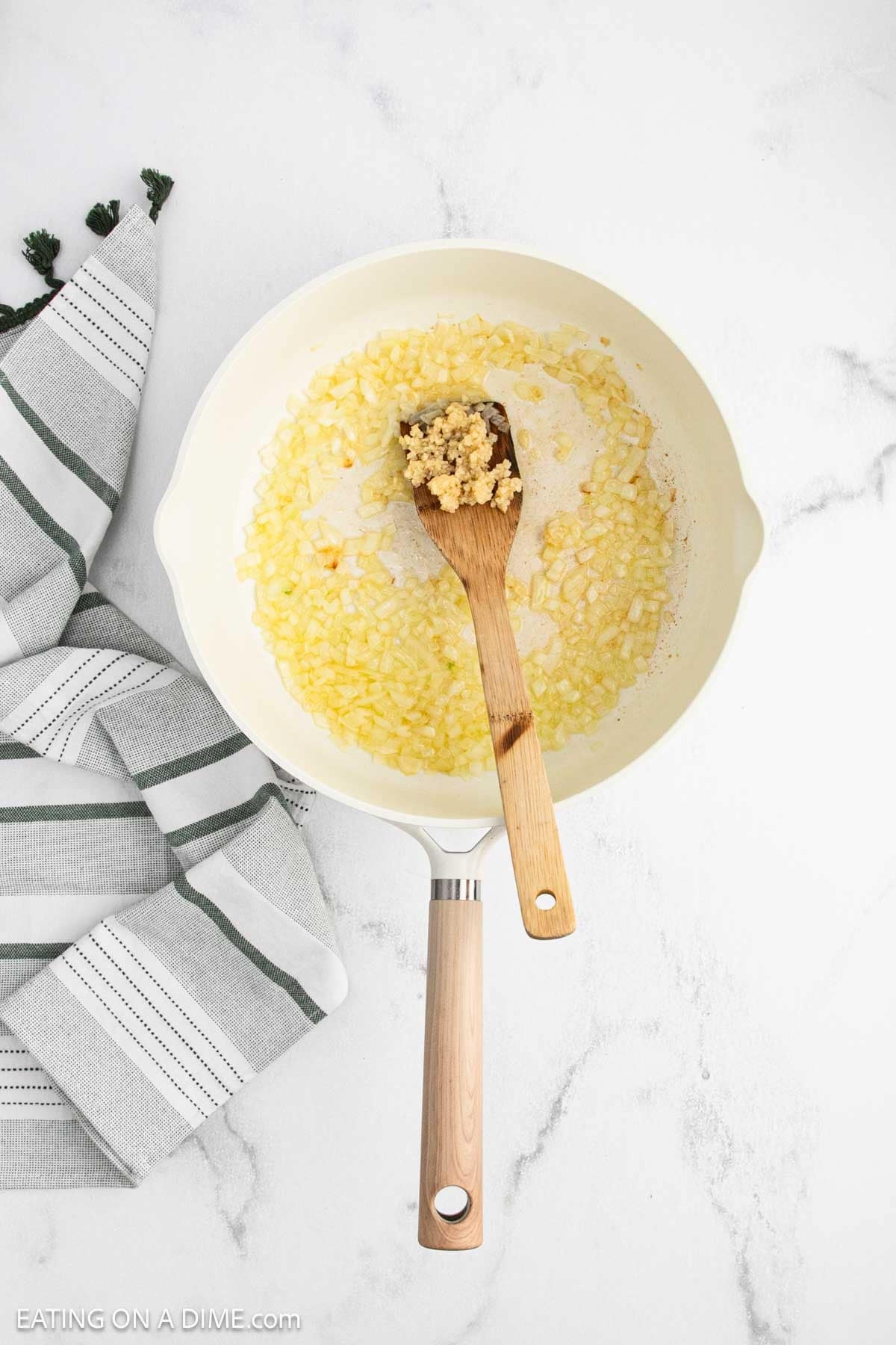 A white skillet with sautéed chopped onions and a wooden spoon holding minced garlic.