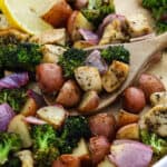 A wooden spoon scoops up Sheet Pan Chicken and Veggies—roasted broccoli, red potatoes, red onions, and seasoned chicken—with a lemon wedge in the background.