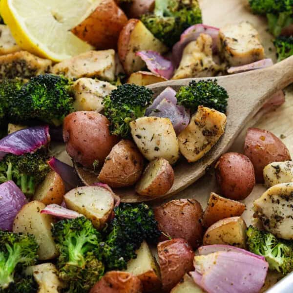 A wooden spoon scoops up Sheet Pan Chicken and Veggies—roasted broccoli, red potatoes, red onions, and seasoned chicken—with a lemon wedge in the background.