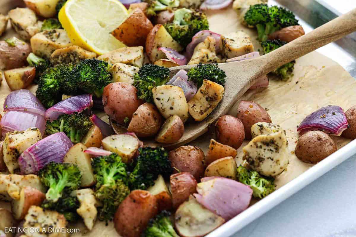 A sheet pan of Sheet Pan Chicken and Veggies features roasted chicken, red potatoes, broccoli, and red onions, garnished with herbs. A wooden spoon serves the mixture while a lemon wedge brightens the corner.