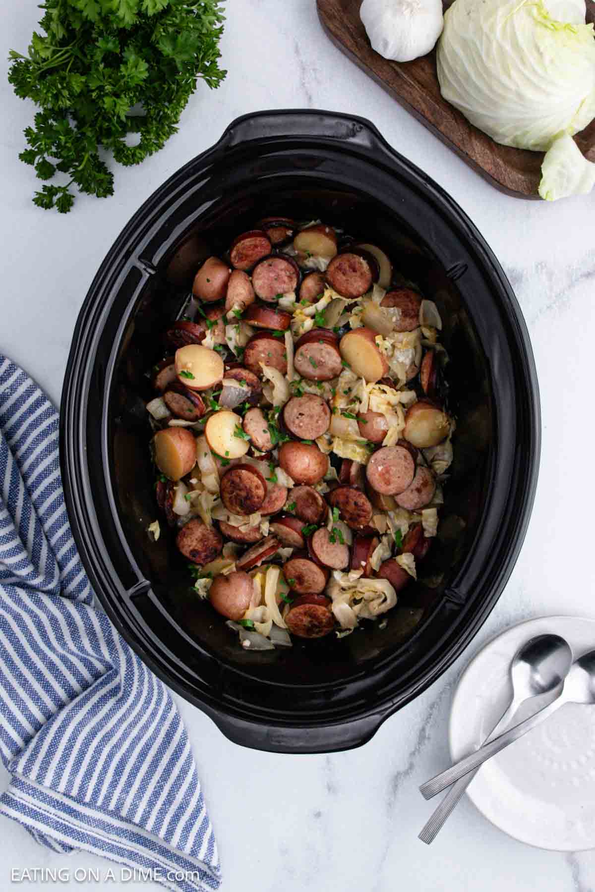 Overhead view of a slow cooker brimming with sausage, potatoes, and cabbage. Nearby are a striped napkin, two stacked white bowls with spoons, fresh parsley, garlic, and a wooden board topped with cabbage.