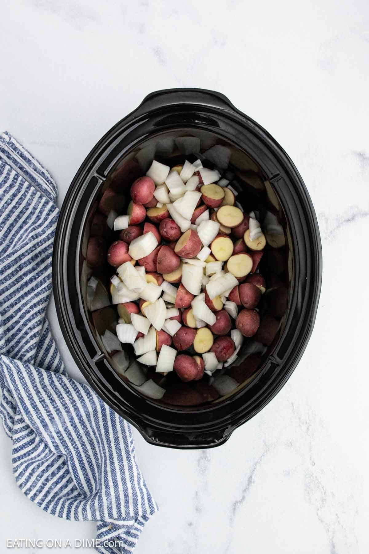 A black slow cooker filled with chopped red potatoes, diced onions, and savory sausage sits on a white marble surface next to a blue and white striped kitchen towel—perfect for hearty cabbage and sausage recipes.