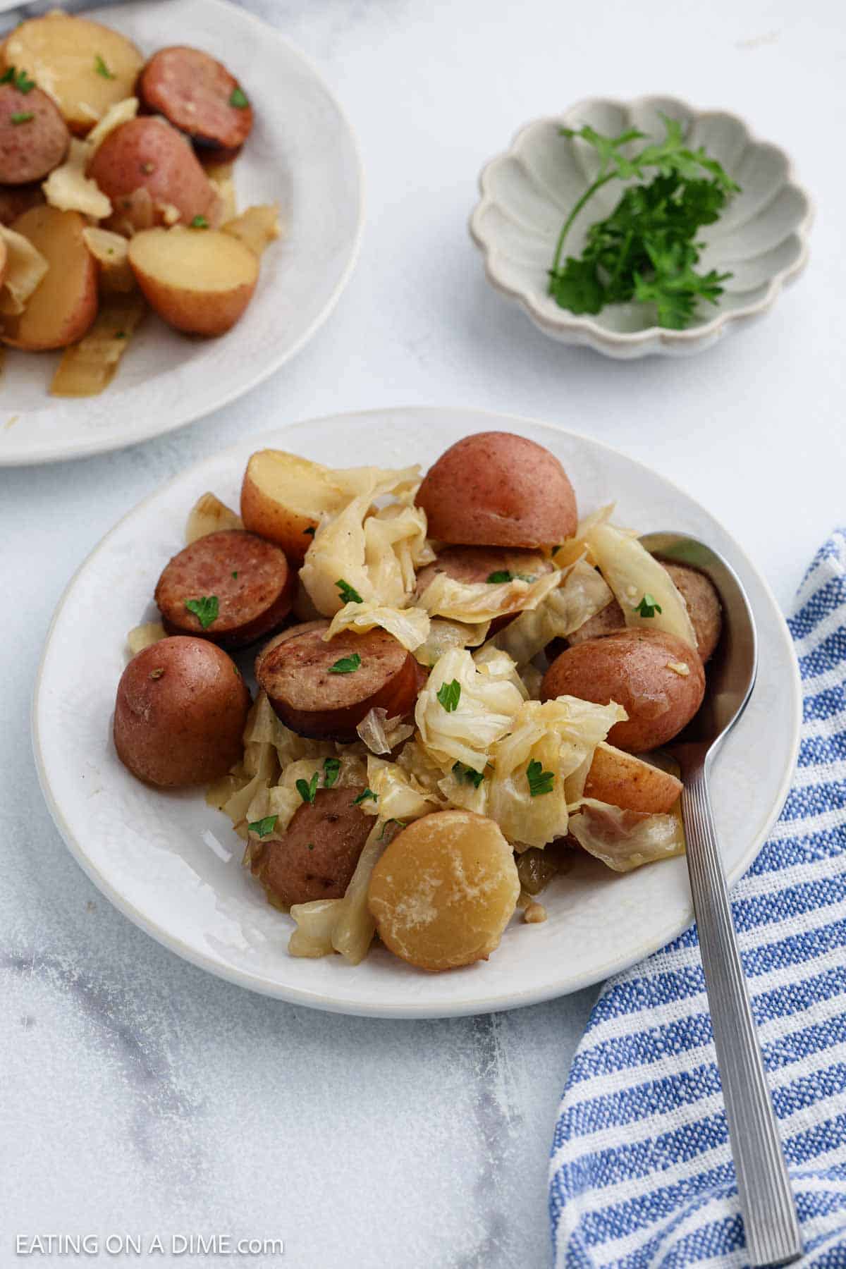 A white plate holds cooked red potatoes, sliced sausage, and tender slow cooker cabbage, garnished with chopped parsley. A spoon rests on the plate. In the background, there's another serving, a small dish with parsley, and a blue-striped napkin.