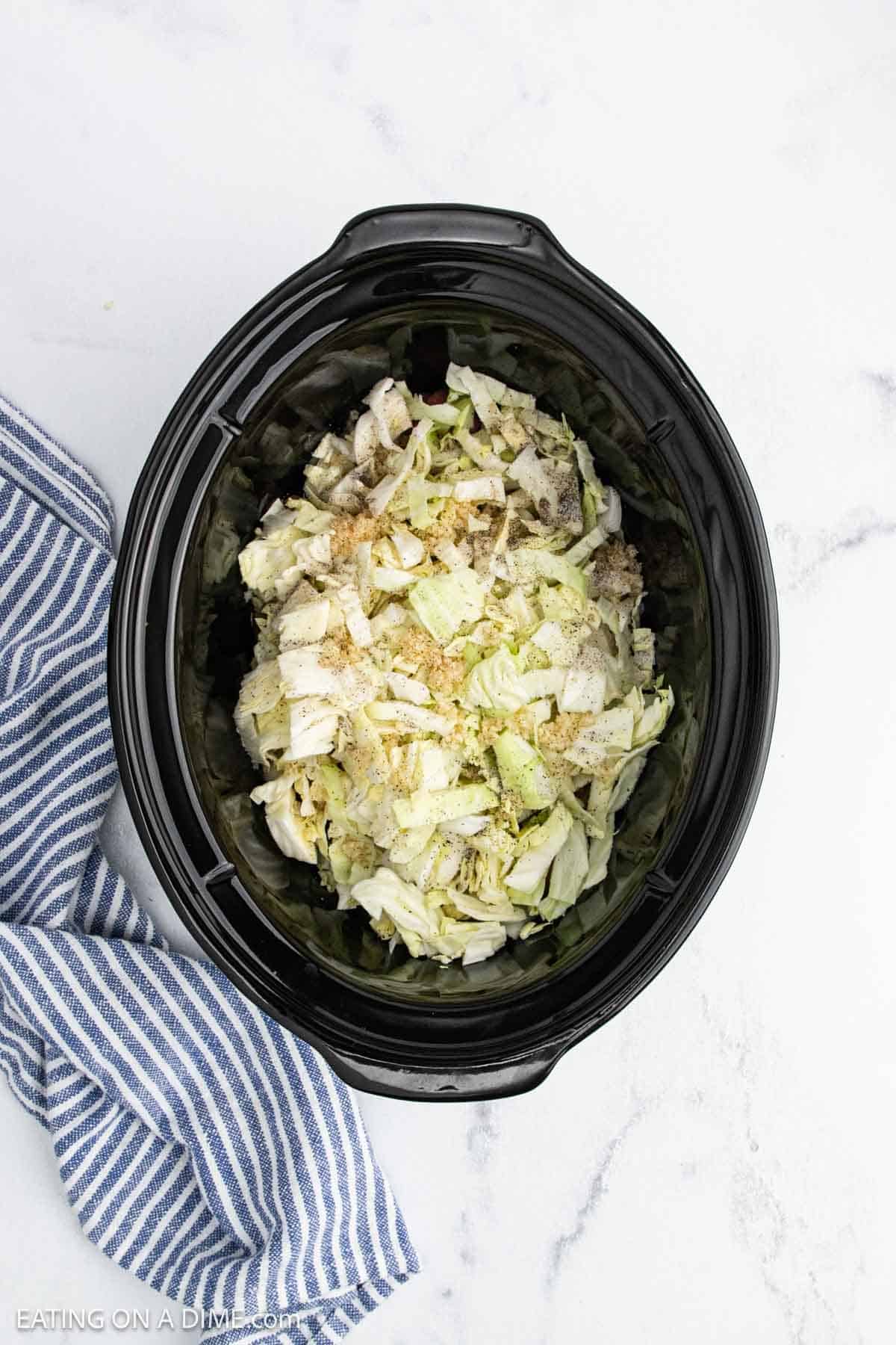 A black slow cooker filled with chopped cabbage and seasonings sits on a white marble surface next to a blue and white striped cloth, perfect for hearty slow cooker cabbage or other comforting slow cooker recipes.