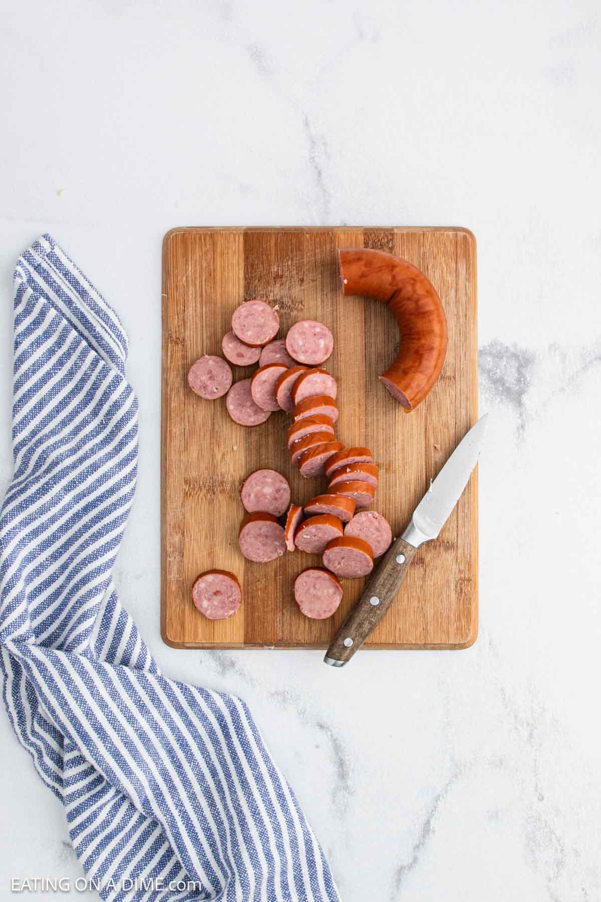 A wooden cutting board with sliced sausage, a knife, and a partially cut sausage—perfect for adding to your favorite slow cooker recipes. A blue and white striped kitchen towel is draped next to the board on a white countertop.