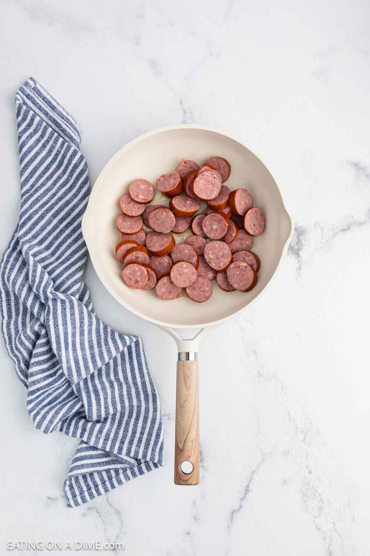 A white skillet filled with sliced sausage and tender cabbage sits on a marble countertop next to a blue and white striped kitchen towel.