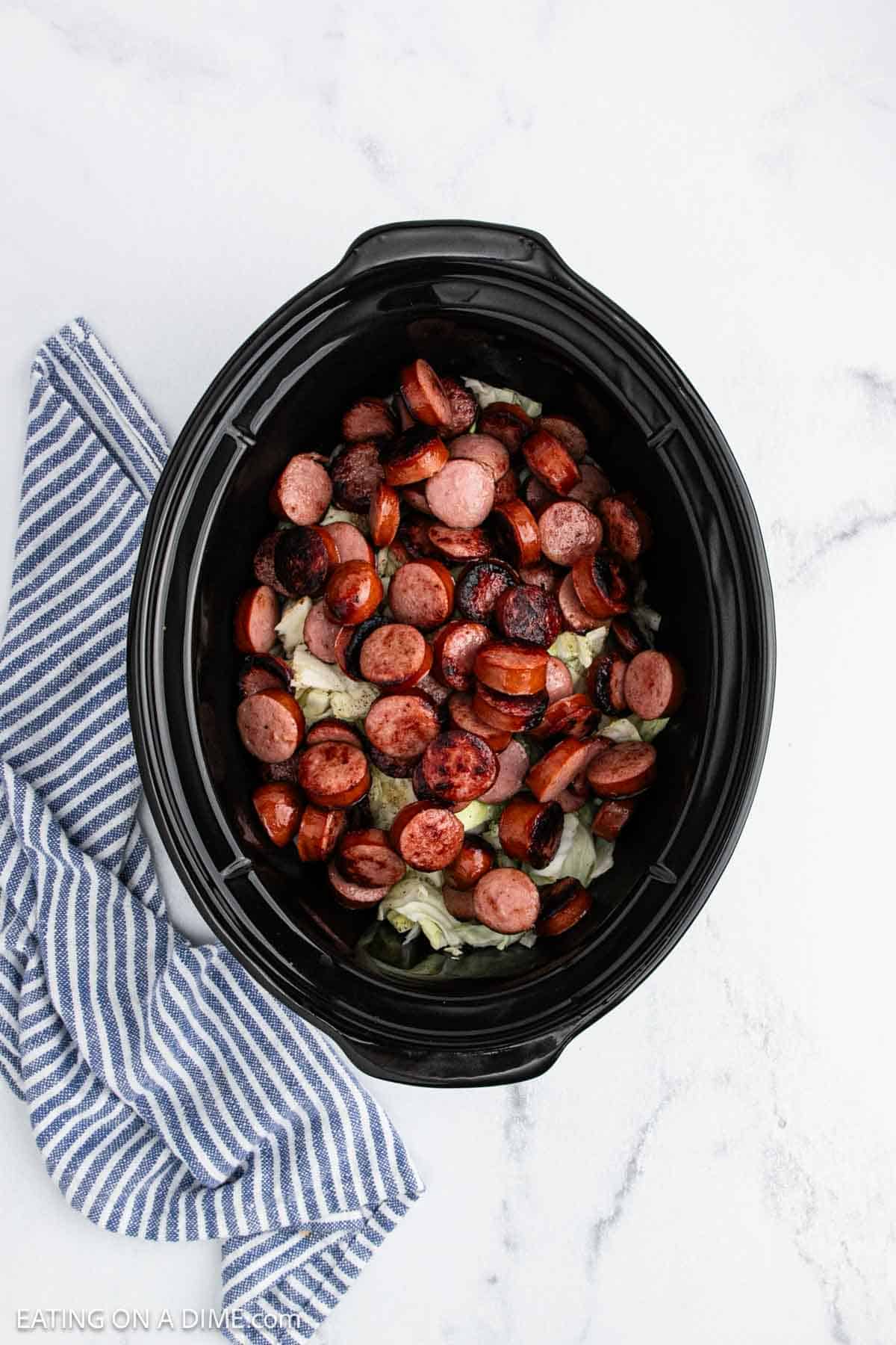 A black slow cooker filled with savory sausage and tender cabbage sits on a white marble surface, next to a blue and white striped kitchen towel.