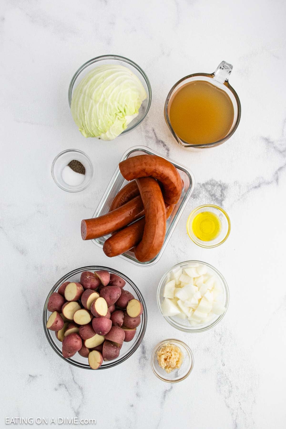Ingredients for a Slow Cooker sausage and cabbage dish arranged on a white surface: sliced cabbage, chicken broth, smoked sausage links, halved red potatoes, chopped onion, minced garlic, olive oil, salt, and pepper.