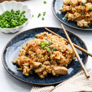 A plate of Teriyaki Chicken Rice with vegetables and green onions, garnished with chopped scallions. A pair of chopsticks rests on the plate, with a bowl of more green onions in the background.