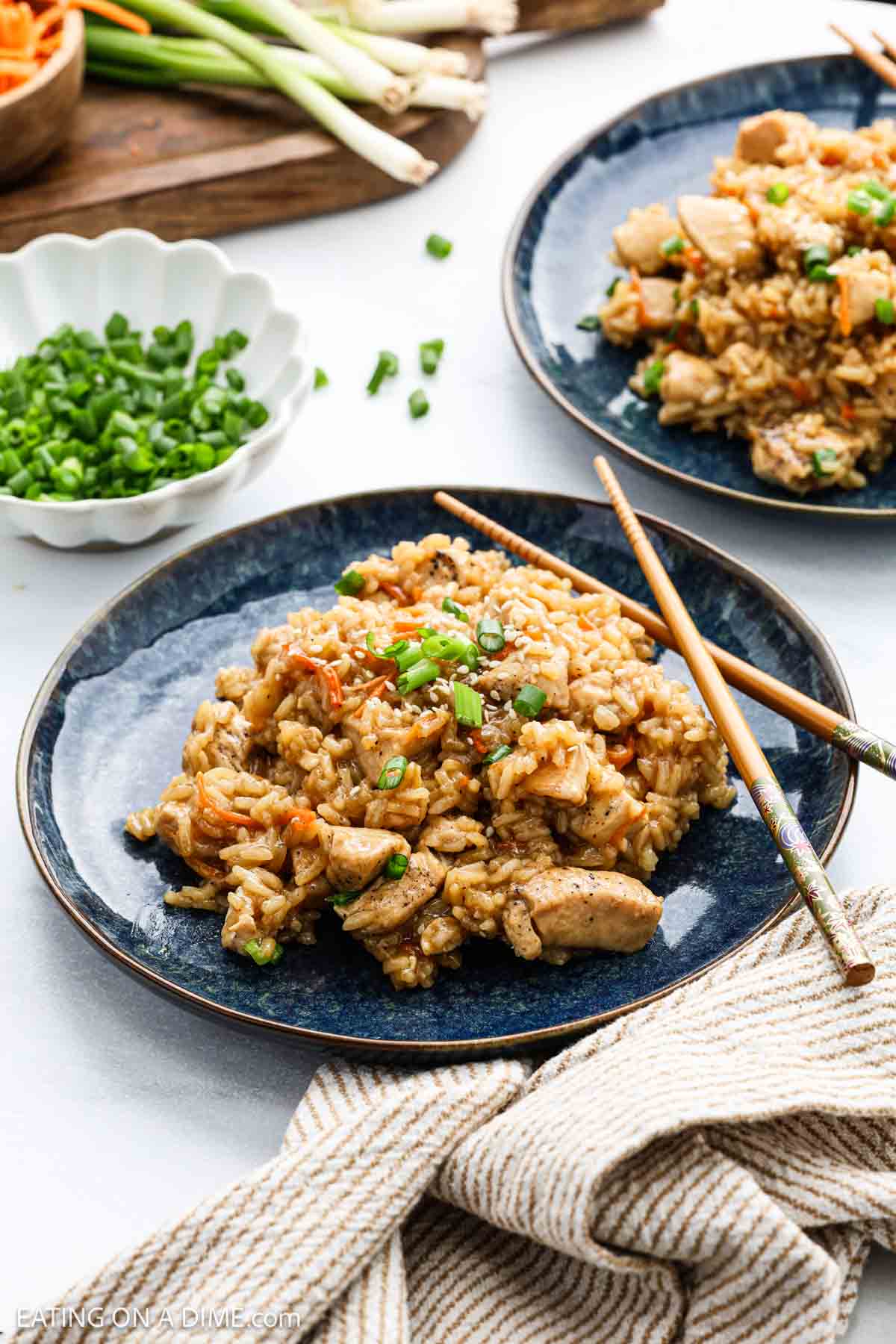 A dark blue plate of Teriyaki Chicken Rice garnished with chopped green onions, with wooden chopsticks resting on the plate. Additional plates of food and bowls of green onions are in the background.