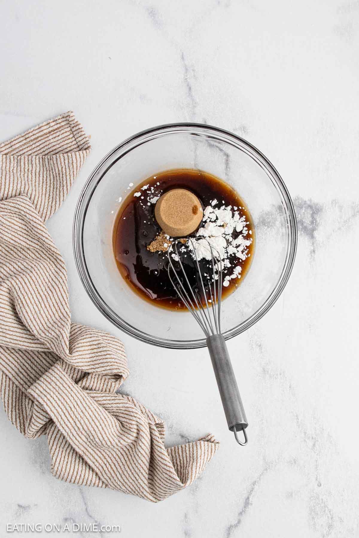 A glass bowl with brown sugar, soy sauce, and cornstarch being whisked together on a marble countertop—perfect for making Teriyaki Chicken—sits next to a beige striped kitchen towel.