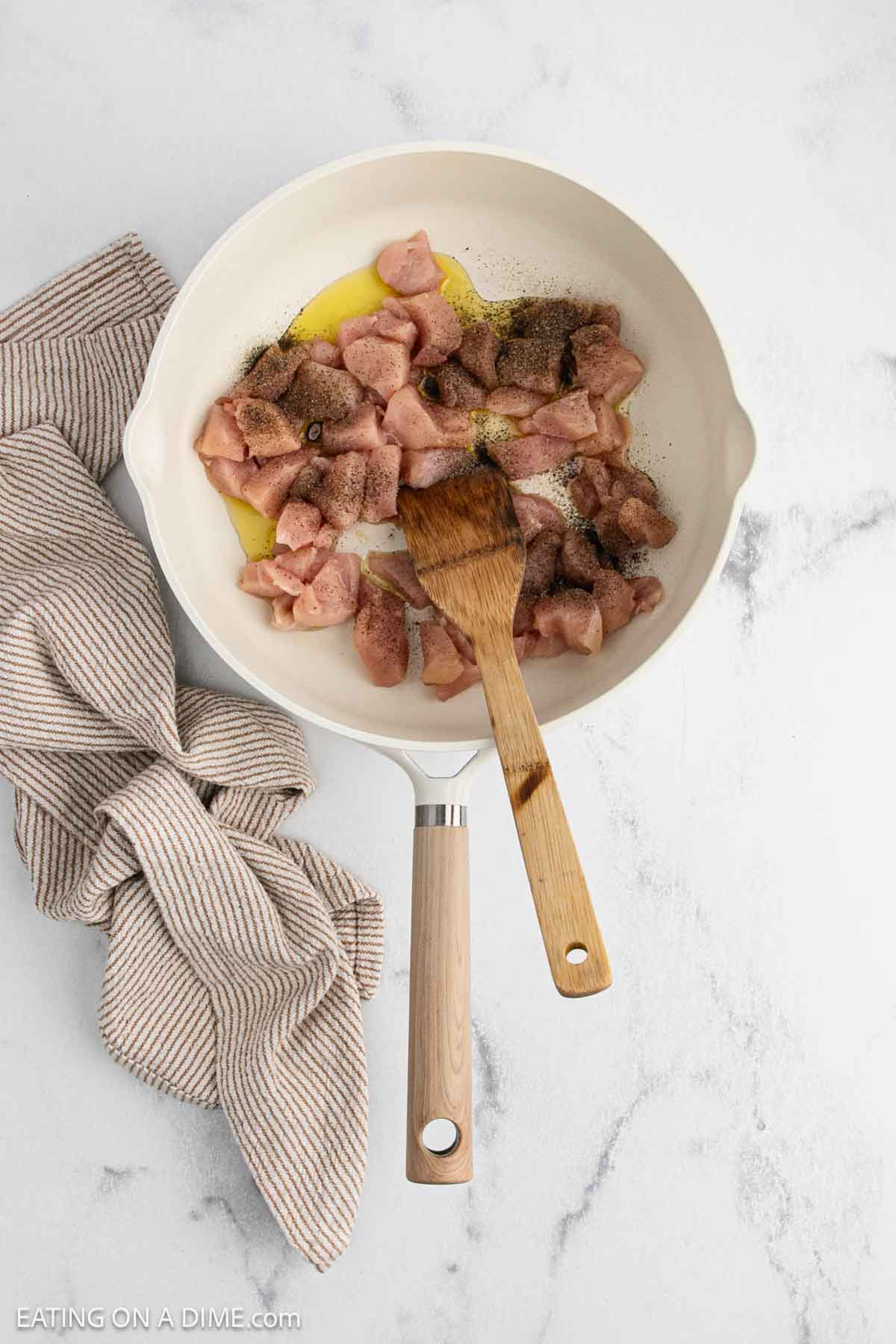 A white skillet with diced raw chicken, oil, and seasoning is being stirred with a wooden spatula—perfect for beginning a flavorful Teriyaki Chicken Rice dish. A striped kitchen towel rests nearby on the white marble surface.