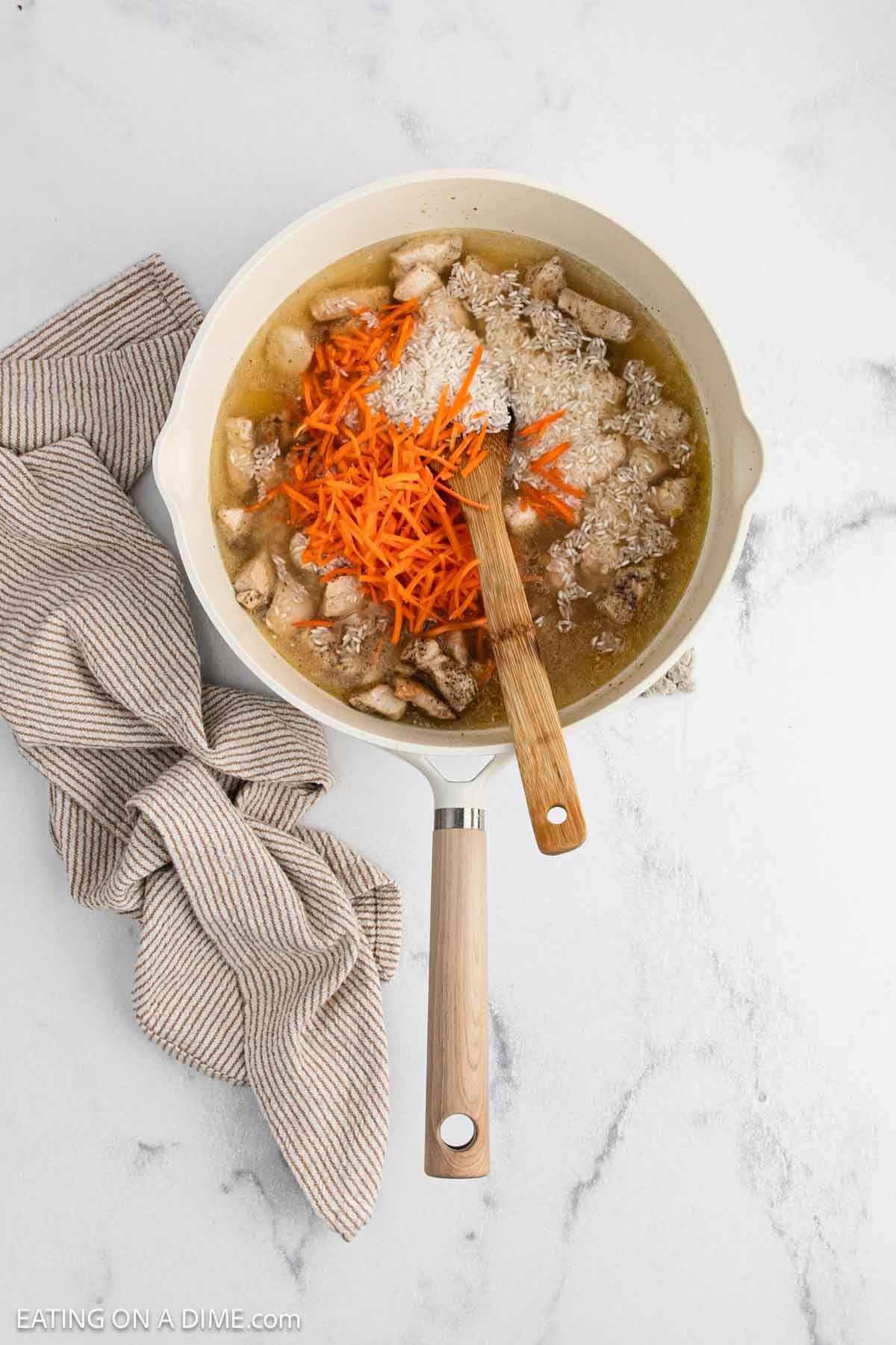 A white skillet of Teriyaki Chicken Rice, with sliced chicken, shredded carrots, uncooked rice, and broth, sits on a marble surface beside a beige striped kitchen towel. A wooden spoon rests in the skillet.