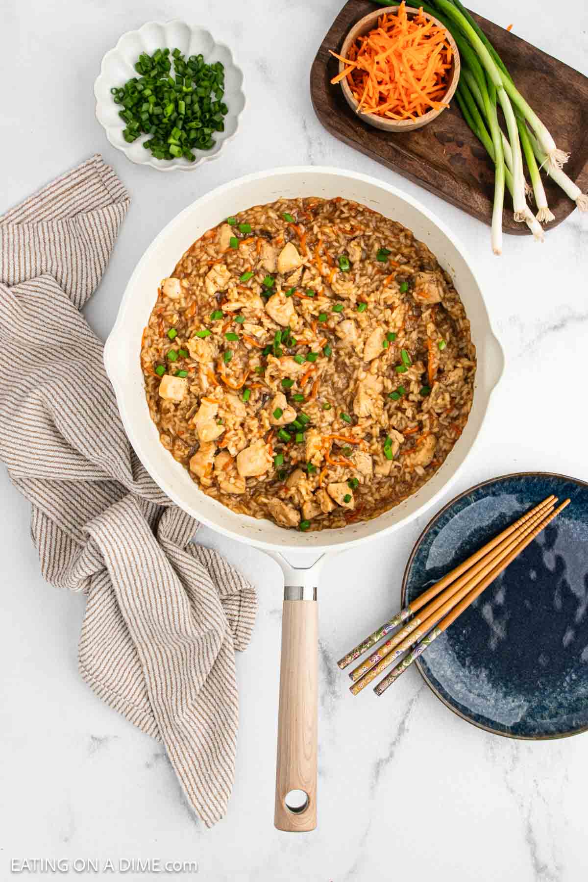 A skillet filled with teriyaki chicken and rice, vegetables, and tender chicken pieces sits on a marble counter, surrounded by a striped towel, shredded carrots, green onions, and plates with chopsticks.