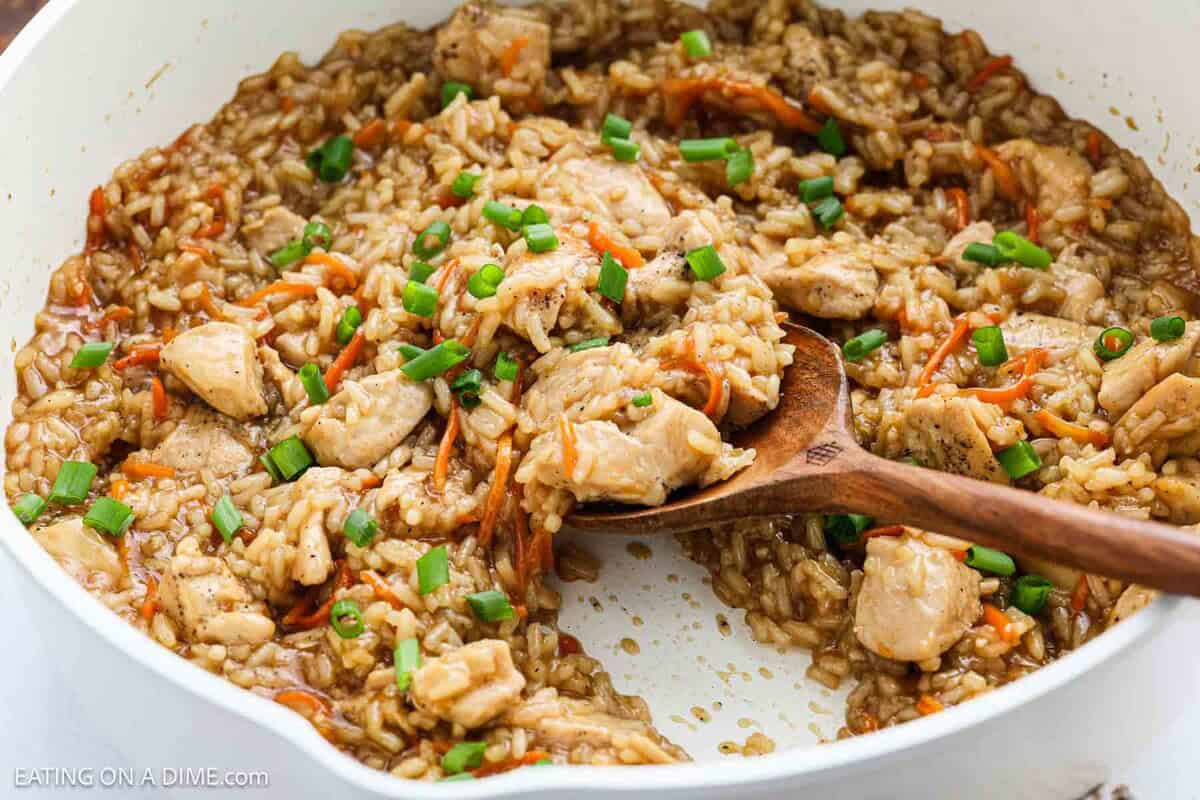 A close-up of a skillet filled with Teriyaki Chicken Rice, garnished with chopped green onions and shredded carrots, with a wooden spoon scooping a portion.