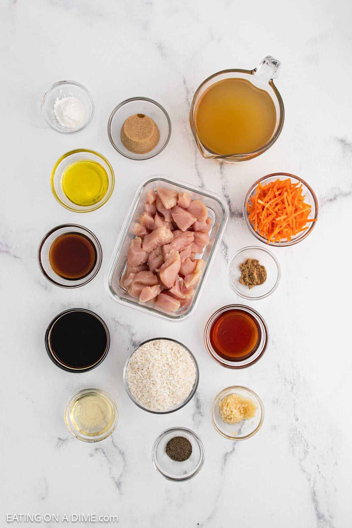 Overhead view of Teriyaki Chicken Rice ingredients in small bowls, featuring diced raw chicken, shredded carrots, rice, chicken broth, sauces, oil, spices, and seasonings—all neatly arranged on a white marble surface.