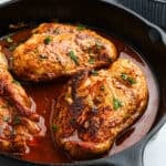 Three seasoned chicken breasts are cooked in a cast iron skillet with a rich, reddish sauce reminiscent of Texas Roadhouse flavors, then garnished with chopped fresh herbs. The chicken is golden-brown and looks juicy and flavorful.