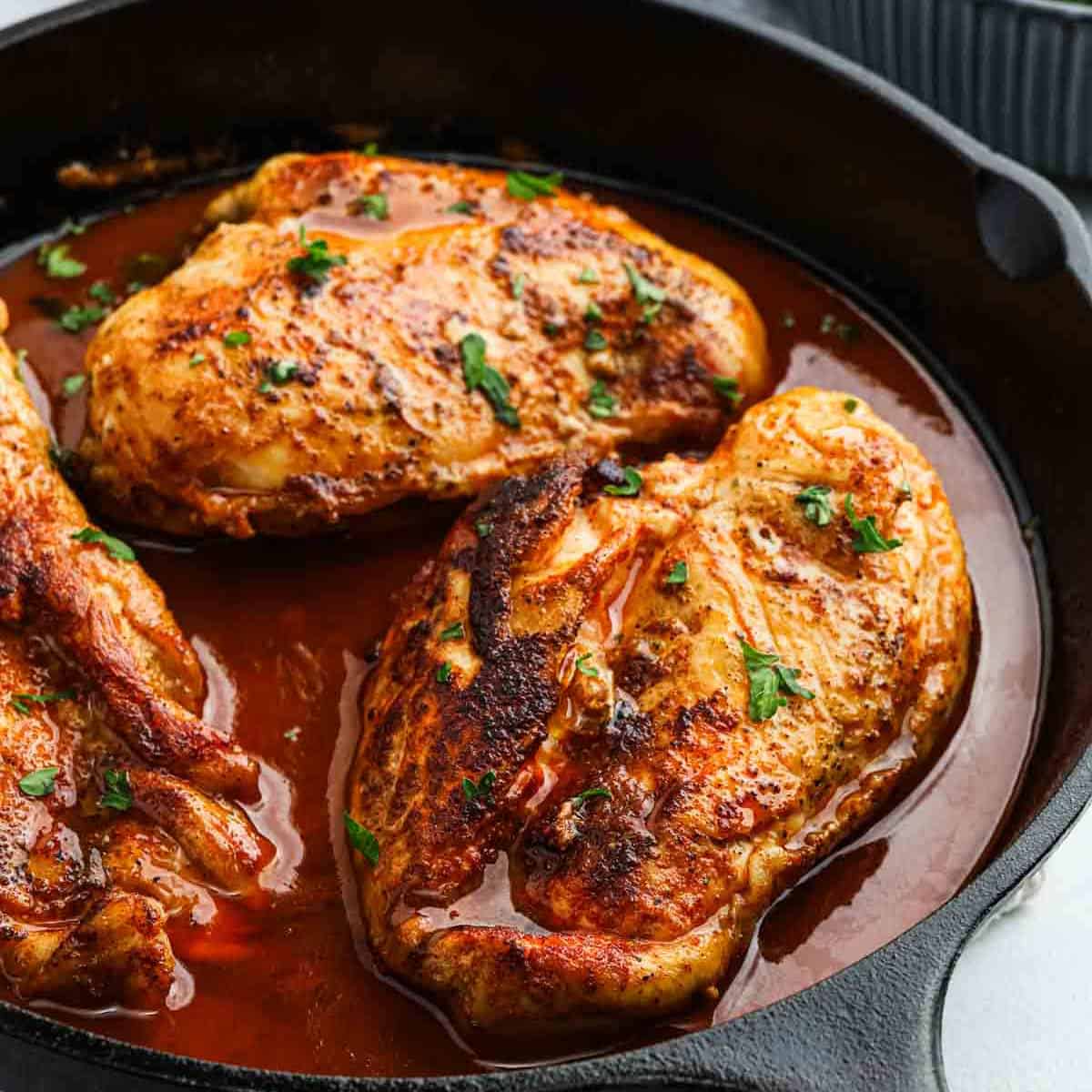 Three seasoned chicken breasts are cooked in a cast iron skillet with a rich, reddish sauce reminiscent of Texas Roadhouse flavors, then garnished with chopped fresh herbs. The chicken is golden-brown and looks juicy and flavorful.