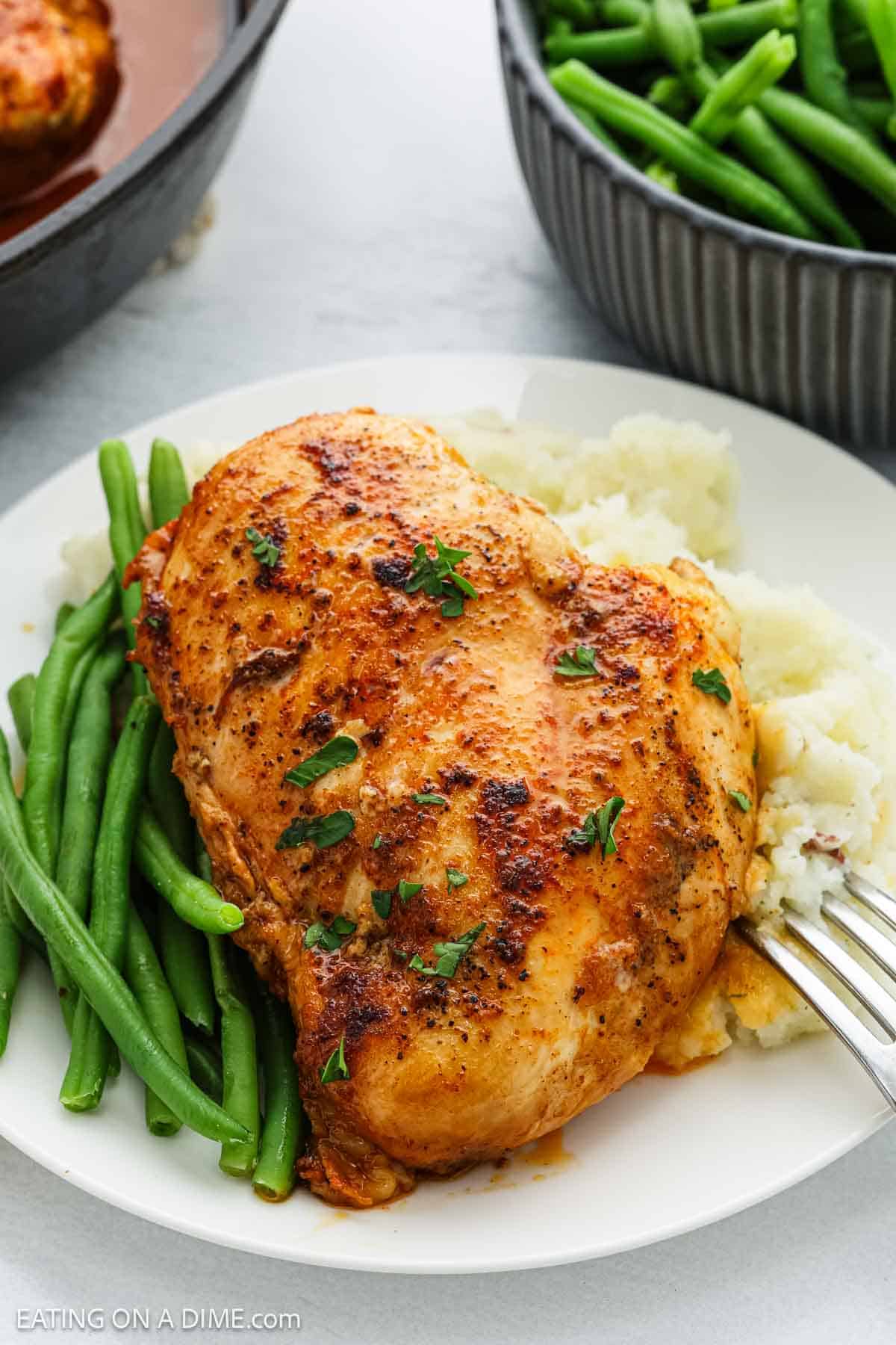 A plate with a seasoned, skillet-cooked chicken breast served on mashed potatoes, garnished with chopped parsley, alongside fresh green beans. A fork rests on the plate and a bowl of green beans is visible in the background.