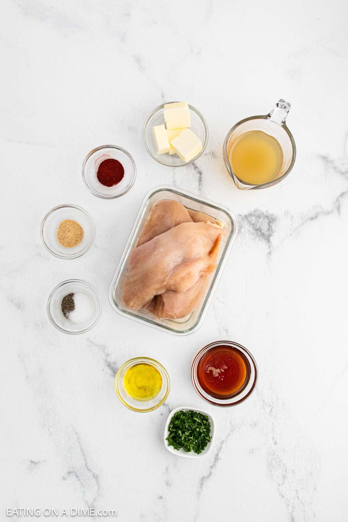 Raw chicken breasts in a glass container surrounded by small bowls of butter, chicken broth, paprika, garlic powder, pepper, olive oil, hot sauce, and chopped parsley—perfect for crafting a Texas Roadhouse-inspired Butter Chicken skillet dish.