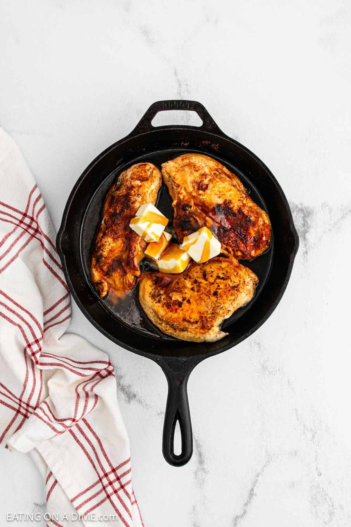 A cast iron skillet with three cooked, seasoned Texas Roadhouse-style chicken breasts and three cubes of butter on top, set on a white marble surface with a red and white striped kitchen towel beside it.