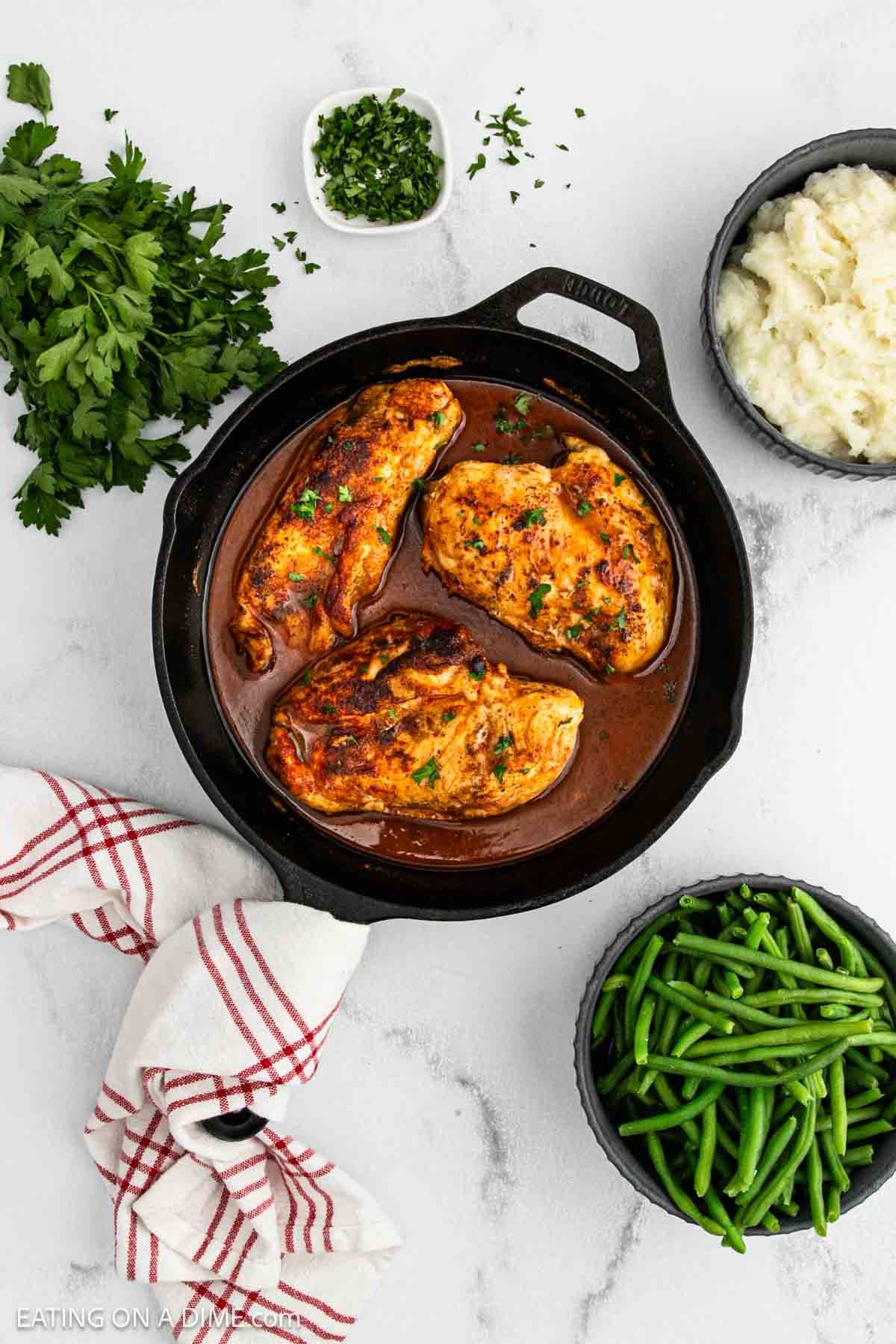 A cast iron skillet with three cooked, seasoned chicken breasts in sauce, reminiscent of Butter Chicken, surrounded by bowls of mashed potatoes, green beans, and chopped herbs on a white surface with a red-striped towel.
