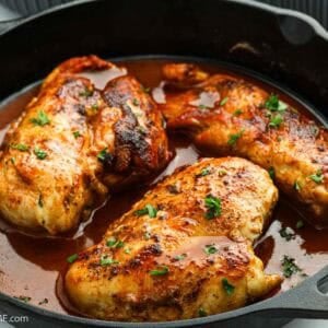 Three cooked chicken breasts in a cast iron skillet, topped with chopped herbs and nestled in a rich brown sauce, reminiscent of a Texas Roadhouse favorite.