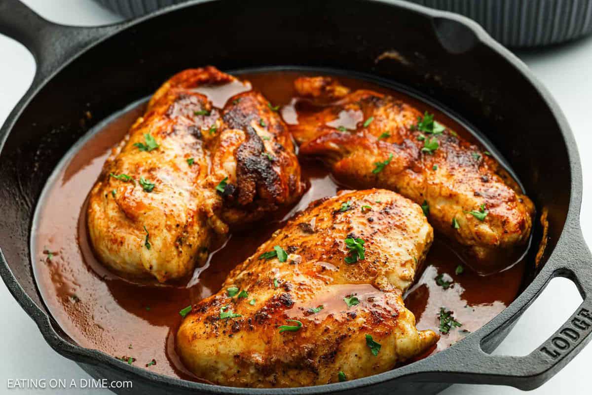 Three cooked chicken breasts in a cast iron skillet, topped with chopped herbs and nestled in a rich brown sauce, reminiscent of a Texas Roadhouse favorite.