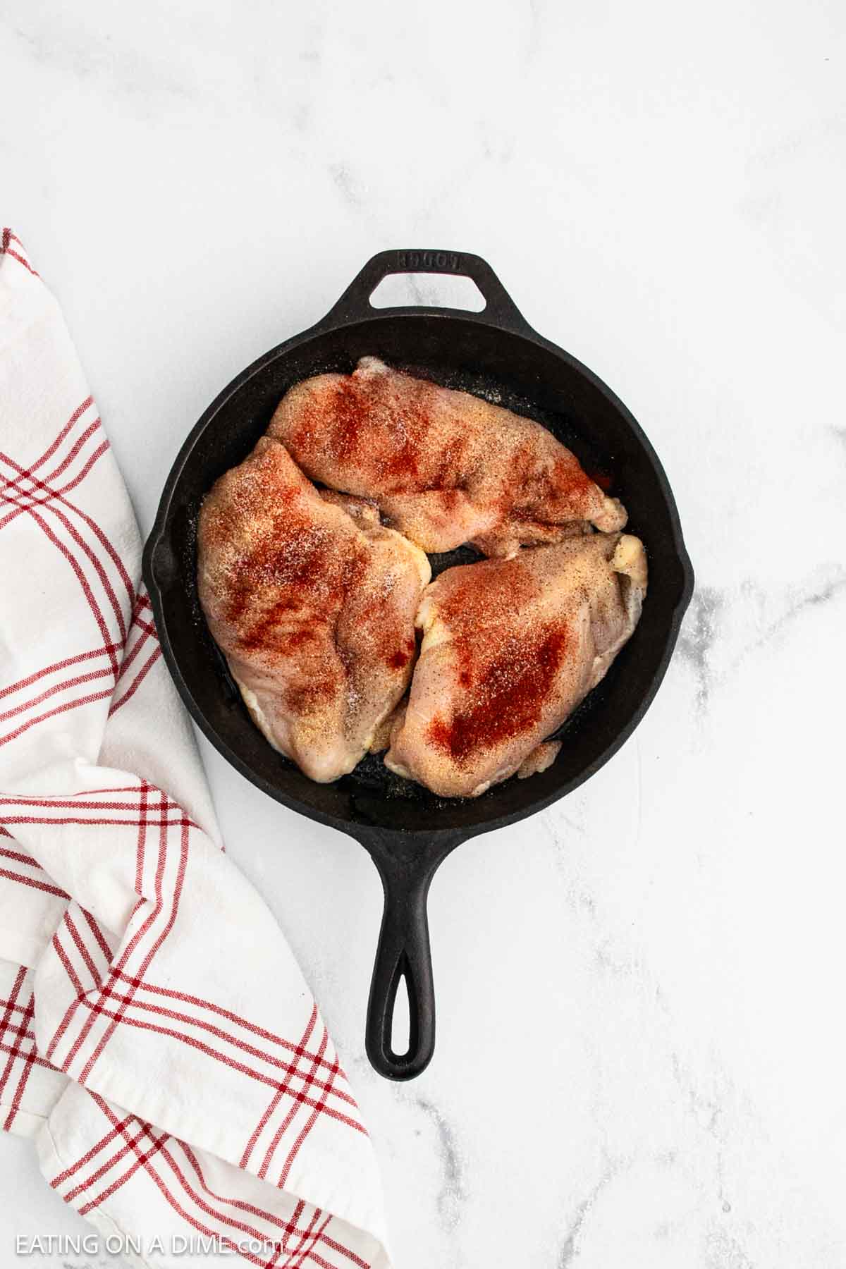 A black cast iron skillet with three raw, seasoned chicken breasts sits on a white marble surface beside a white and red striped kitchen towel—perfect for starting homemade Texas Roadhouse-inspired Butter Chicken.