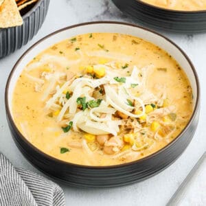A bowl of creamy White Chicken Chili topped with shredded cheese, corn, beans, and chopped herbs sits on a light surface next to a bowl of tortilla chips and a striped napkin.