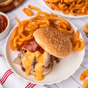 A classic Arby's Beef and Cheddar sandwich with roast beef, cheese, and barbecue sauce on a sesame seed bun, served with curly fries on a white plate. A bowl of sauce and more curly fries complete this Arbyโs meal in the background.